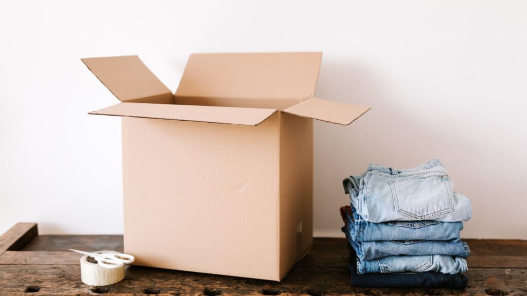 Stacked modern jeans placed near opened cardboard box and tap with scissors before packing for moving out in daylight
