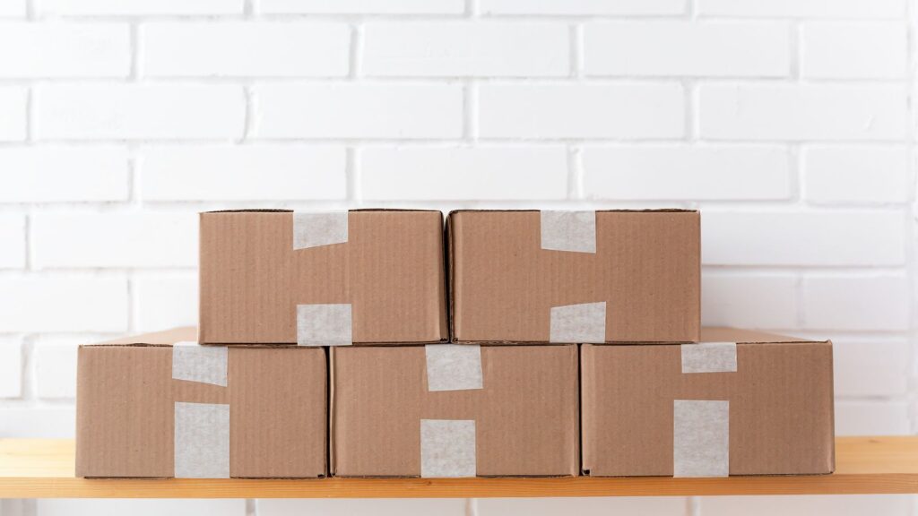 A neat stack of cardboard boxes taped and placed on a wooden shelf against a white brick wall.