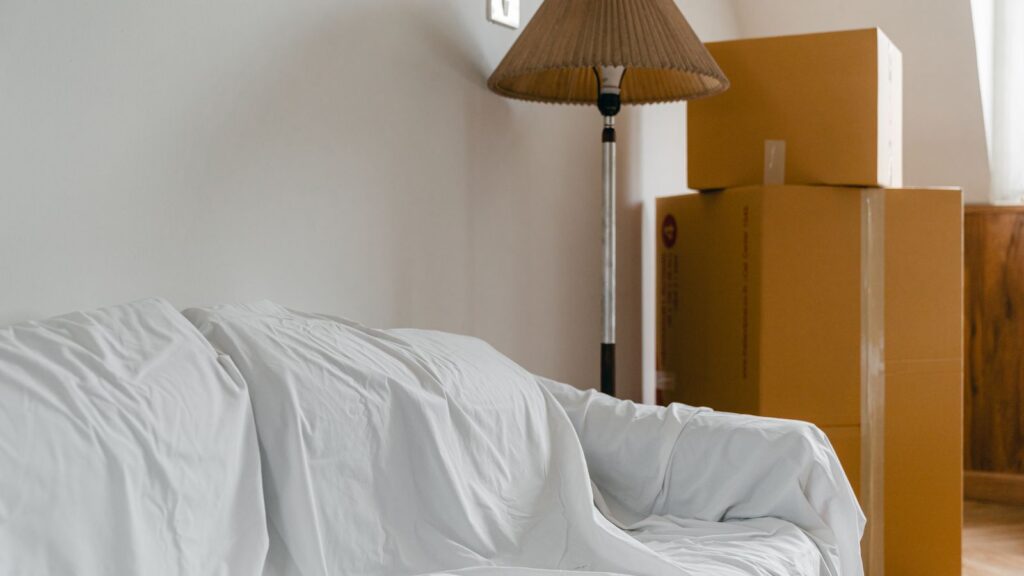 Covered with sheet sofa and floor lamp and packed cardboard boxes in light spacious room of new contemporary house